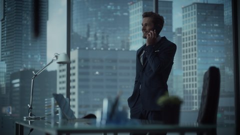Charismatic Young Businessman in a Suit Standing in Modern Office, Talking on a Phone, Looking out of the Window on Big City with Skyscrapers. Successful Finance Manager Planning Work Projects.