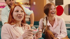 A close up of a ginger haired woman winning in video games while other five people around her are watching her play and sitting in blue bean bag chairs