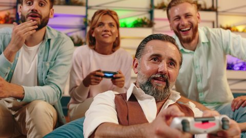 A close up of an older man with grey hair playing against a younger blonde woman in video games and the blonde woman winning, everyone else around them cheering excitedly
