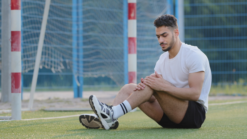 Unhappy injured Middle Eastern indian footballer sitting on grass of soccer field against goal, holding knee. Arabian hispanic athlete in sportswear suffers from leg pain experiencing about injury Royalty-Free Stock Footage #1079839670