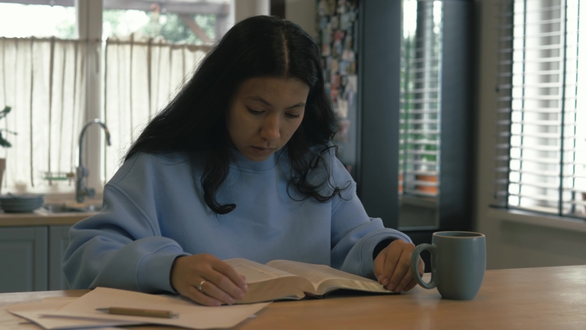 Young female student study at home in the morning drinking tea or coffee. Christian hispanic girl with black hair reading holy book, bible. Devotional | Shutterstock HD Video #1079841848