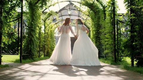 TRACKING Candid shot of two happy lesbian LGBT brides wearing boho dresses walking through alley on their wedding day. Shot with 2x anamorphic lens