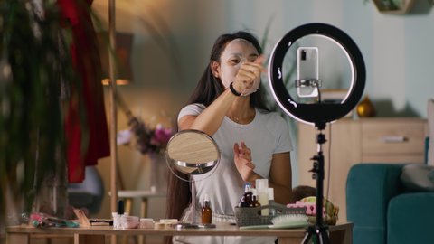 Asian woman in moisturizing sheet mask asking audience to like video while sitting at table near child and recording beauty vlog at home