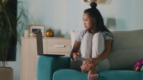 Young Asian woman filing toenails and watching video on netbook while sitting on couch and filing nails doing pedicure at home