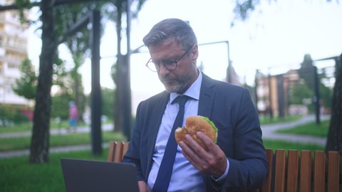 Man in suit eating burger while working on laptop outdoor, feeling stomach pain