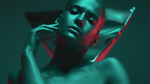 Portrait of young woman with shiny makeup and skin in colorful neon light. Female model moving sensually looking at camera in studio. Futuristic advertisement of modern fashion