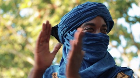 Errachidia, Morocco:29-09-21:Man in traditional blue Tuareg dresses, sitting in a park listening to music and clapping with his hands. Blurred background of trees. 