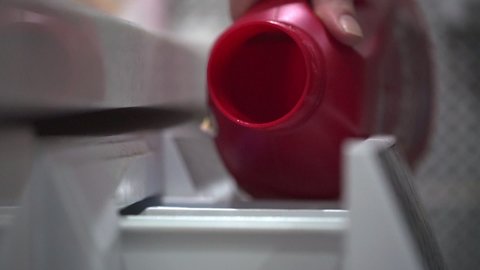 Woman pours necessary amount of white liquid laundry detergent into compartment of washing machine from big red bottle