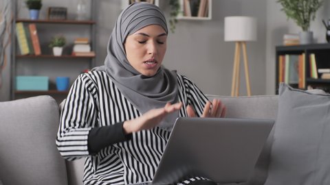 furious stressed muslim woman working at the laptop has a burst of anger,hits her hands on the keyboard,overwhelmed angry arab female sits on couch using computer has a rage attack