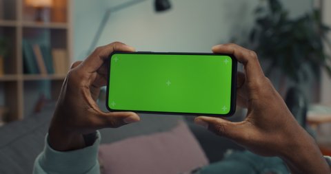 African american guy holding smartphone in horizontal position with green mock up screen. Focus on modern gadgets in male hands. Copy space.