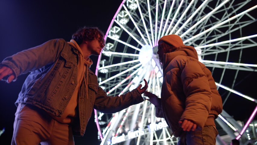 Happy couple playing patty cake game against illuminated ferris wheel at night. Smiling woman and man having fun on urban street. Cheerful girlfriend and boyfriend enjoying time together outdoor. Royalty-Free Stock Footage #1079874620