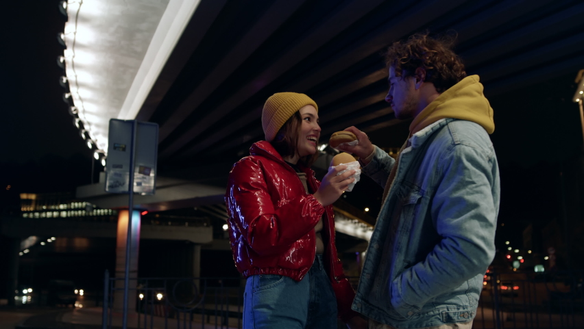Positive couple eating burgers under bridge at night. Happy man feeding woman during romantic date on urban background. Cheerful young pair having snacks food outdoor.  | Shutterstock HD Video #1079874650