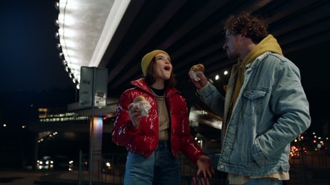 Positive couple eating burgers under bridge at night. Happy man feeding woman during romantic date on urban background. Cheerful young pair having snacks food outdoor. 