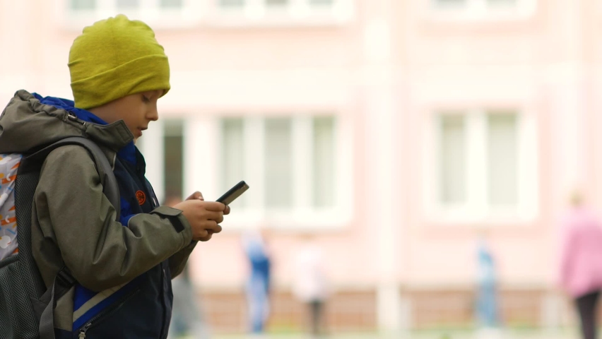 Caucasian Schoolboy 7-9 years old being at the school building plays games on a smartphone. Addiction of children to games. Schoolboy is typing a message on the phone. Children and cyberspace theme.