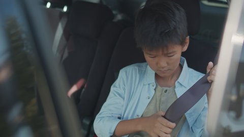 Handheld shot of 12-year-old Asian boy with backpack getting into passenger seat of car and fastening his seatbelt after being picked up from school by his parent or caretaker