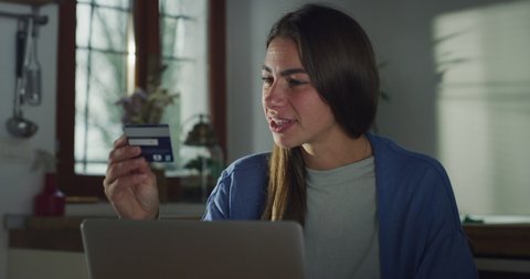 Cinematic shot of young happy smiling woman is entering credit card information by using laptop while is making shopping online in kitchen at home.