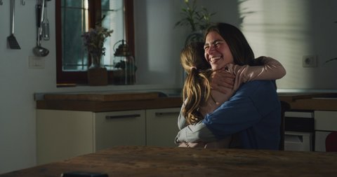 Cinematic authentic shot of happy smiling little daughter is giving affective hug to her mother while enjoying their time together in kitchen at home.