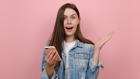 Happy young brunette woman hold using mobile cell phone type say wow yes just found out great big win news doing winner gesture, dressed in denim jacket, isolated on pastel pink color background wall