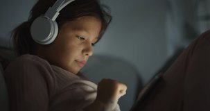 Close Up of a Little Girl Using Headphones and Digital Tablet to Play Games Before Sleeping. Female Kid Focused on a Screen at Night, Watching Videos and Laughing, Having Fun