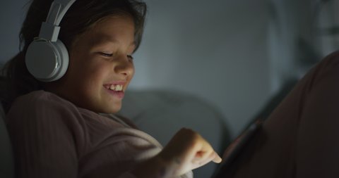 Cinematic shot of happy smiling little girl with headphones is having fun to use technology tablet for family entertainment or online gaming while comfortably sitting on sofa at home in evening.