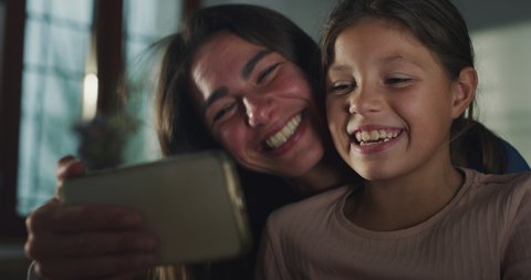 Cinematic close up shot of happy smiling mother and little daughter making selfie and technology video call together with smart phone to father or friends in kitchen at home.