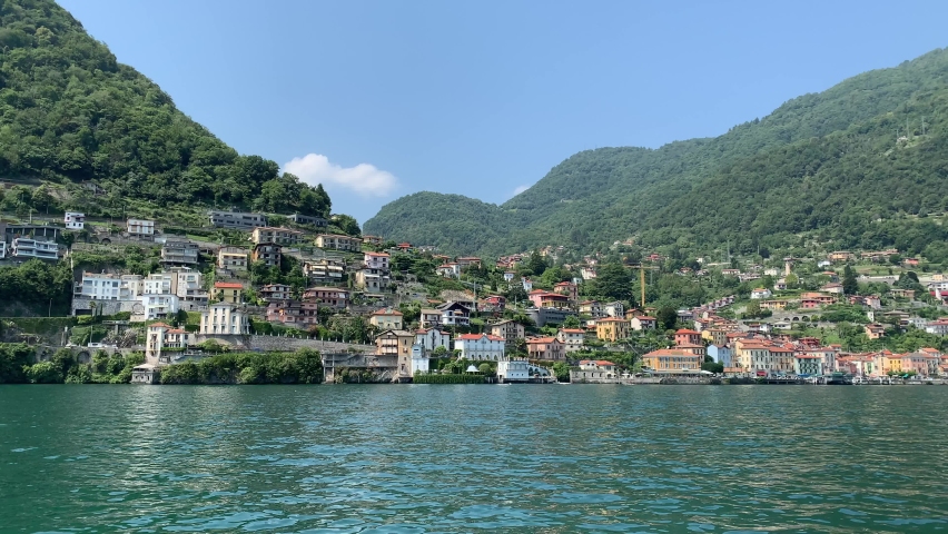 Skyline of Argegno town with houses on hills, mountains. Landscape of Como Lake (Lago di Como) shore. View from the moving boat. Motion of waves and emerald water. Argegno, Como Lake, Lombardy, Italy. | Shutterstock HD Video #1079887859