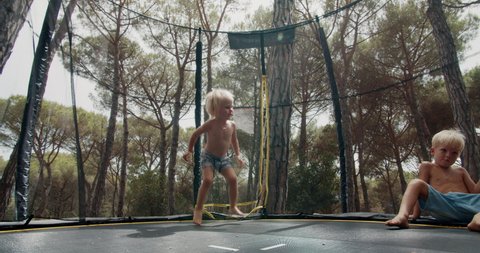 Happy Children baby girl is having fun jumping on trampoline outdoors during summer vacations in Spain. Family spending time together, sister is jumping and brother waiting his turn in forest