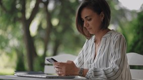 young hispanic latin ethnic woman sitting in a summer cafe holding phone looking at screen watching social media video content, movie or stream online in mobile app laughing using smart phone