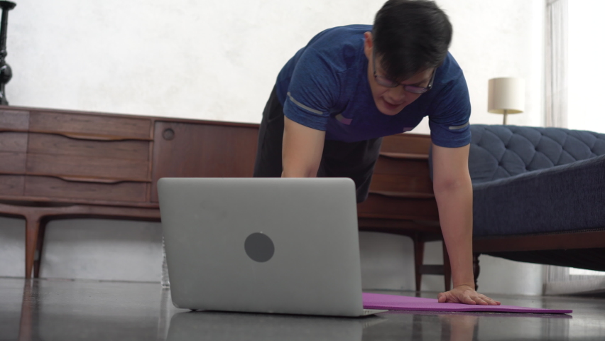 young asian fit man in sportswear doing push ups While watching video training fitness  online tutorials on laptop in living room at home . sport male workout in quarantine 