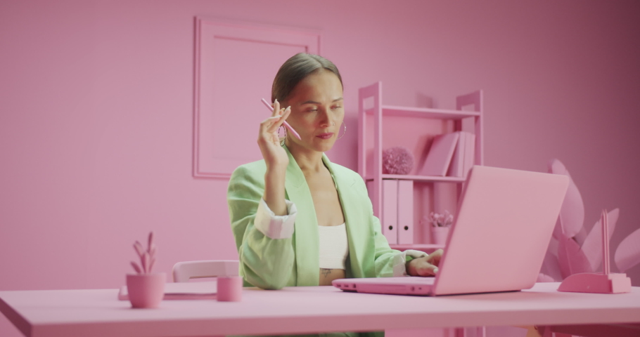 Thoughtful pretty successful female working on project at stylish pink design studio, pensive attractive young Caucasian woman holding pencil thinking about job achievement using laptop on the table Royalty-Free Stock Footage #1079906210