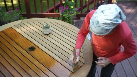 Berkeley Springs, WV, USA - 09 06 2021: Overhead view of mature woman applying varnish to teak table furniture in garden area at home.