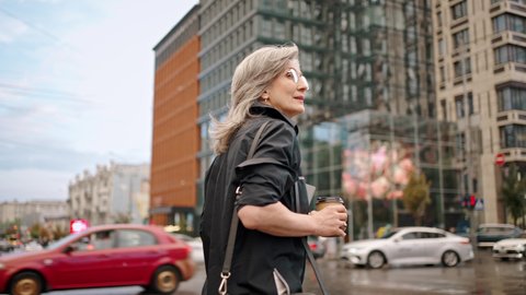Back view. Slow motion of an adult gray-haired woman in glasses walks along a city street with a folder and coffee in her hands. Business lady walk. Slow motion.