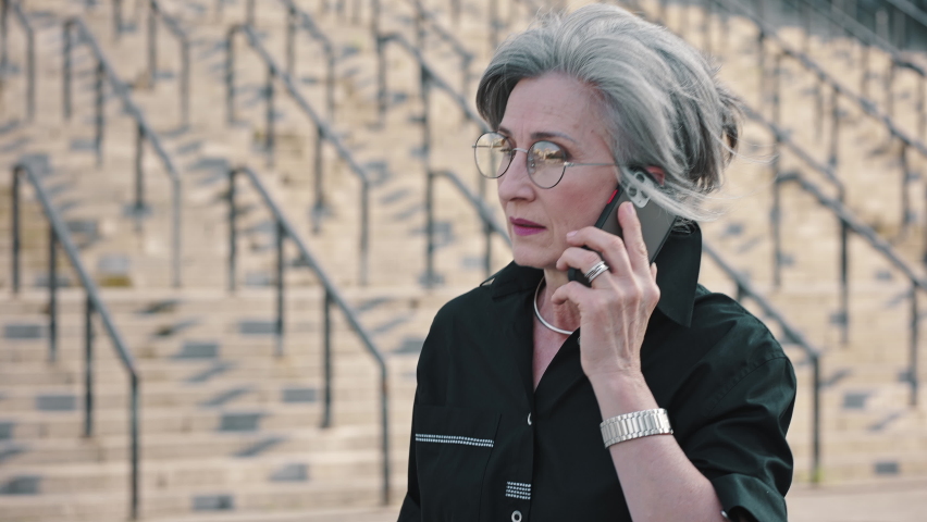 Gray-haired mature lady in black shirt talking on the phone during lunch break. Beautiful senior woman walking outside of business center talking on phone. Slow motion.