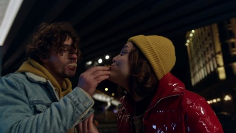 Joyful couple feeding each other on city background at night. Cheerful man and woman enjoying snack food on urban street. Happy girlfriend and boyfriend eating outdoor. Romantic date concept.