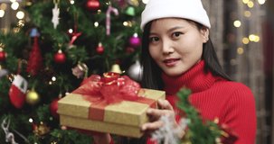 Young teen cute Chinese girl holding present box offer it gives to camera in room with Christmas tree and ornaments, boke lights in background. Idea of Xmas blessing new normal life.