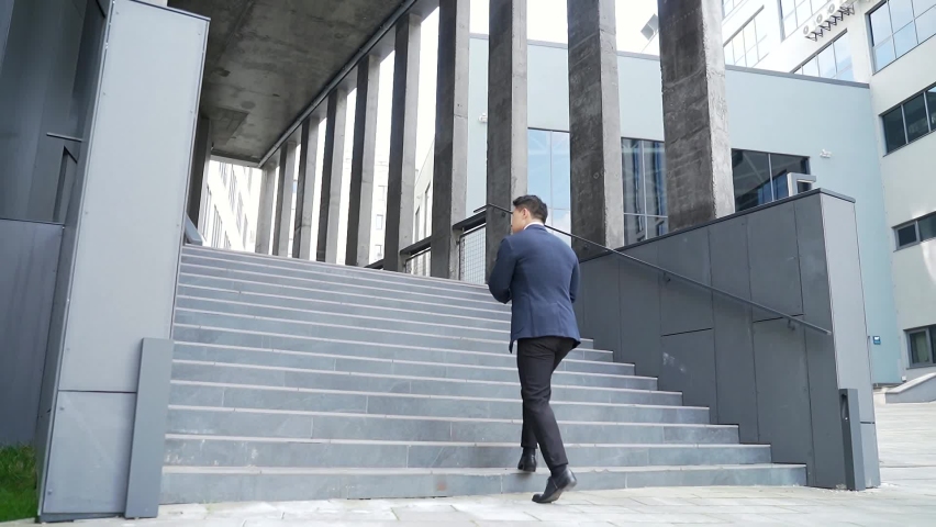 Young successful and confident asian business man fast climbs the stairs up. going modern office building outside. Rear view back. Male businessman executive in formal suit goes to meeting in downtown