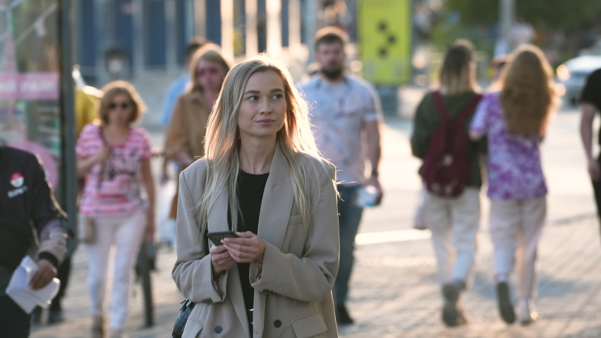 Charming businesswoman, casual looking trendy lady texts on mobile phone. Natural blond woman typing messages on mobile phone touch screen on crowded streets background. Portrait of young adult lady.