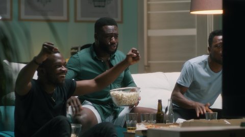 Black men with popcorn screaming and giving high five then clinking glasses and bottles of beer while sitting on couch and watching match on TV at home