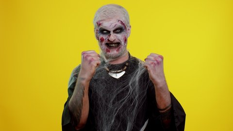 Zombie man with makeup with fake wounds scars celebrate success win scream rejoices doing winner hands gesture say Yes in yellow studio room. Sinister dead guy. Halloween, filming, staging concept