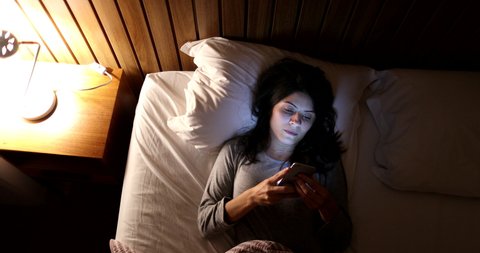 Woman lying in bed turns off smartphone and bedside nightstand lamp