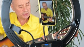 charming elderly man in a yellow shirt plays guitar in front of a ring light and a smartphone, teaches students online skills, records a training video, concept performance, Home Recording Studio