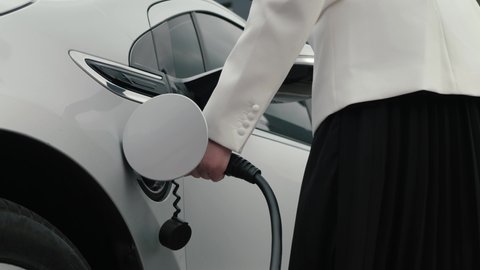 Business woman connects plug to electric car, activates charging app on phone. Eco friendly alternative energy green environment concept. Lady plugs power cable to charge electric car in parking lot