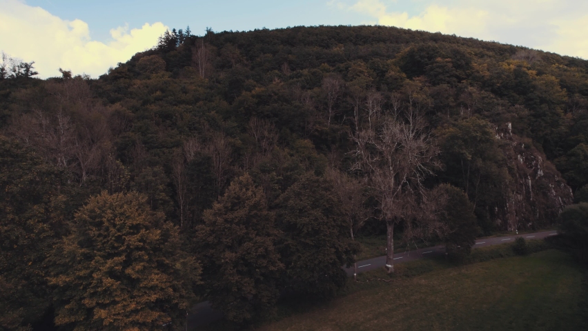 Drone descending in a valley at dusk over grasfield with forests and a road, a  by and a steep rocky mountain in the background. Luxembourg Drone aerial footage
