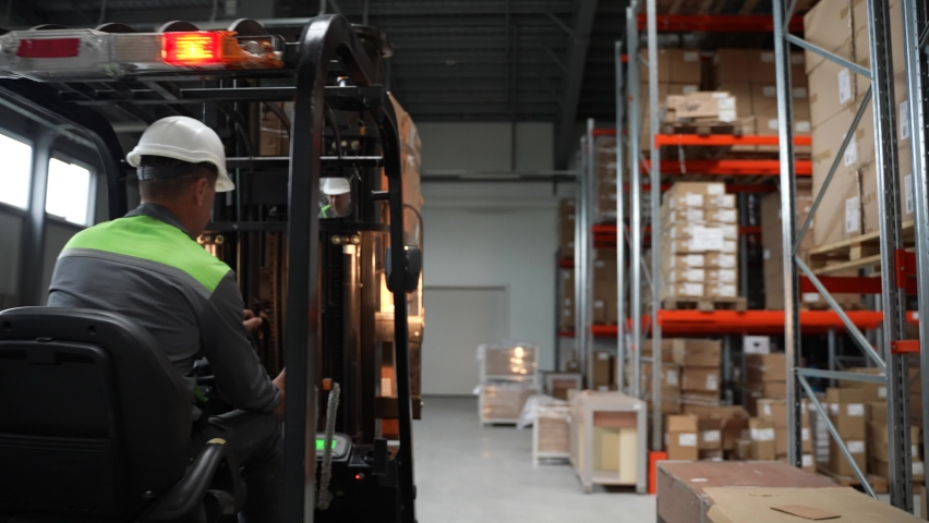 Back view of forklift driver riding between racks in storehouse with pallet of cardboard boxes to place on shelf. Male employee in helmet driving warehouse loader during work Royalty-Free Stock Footage #1079954765