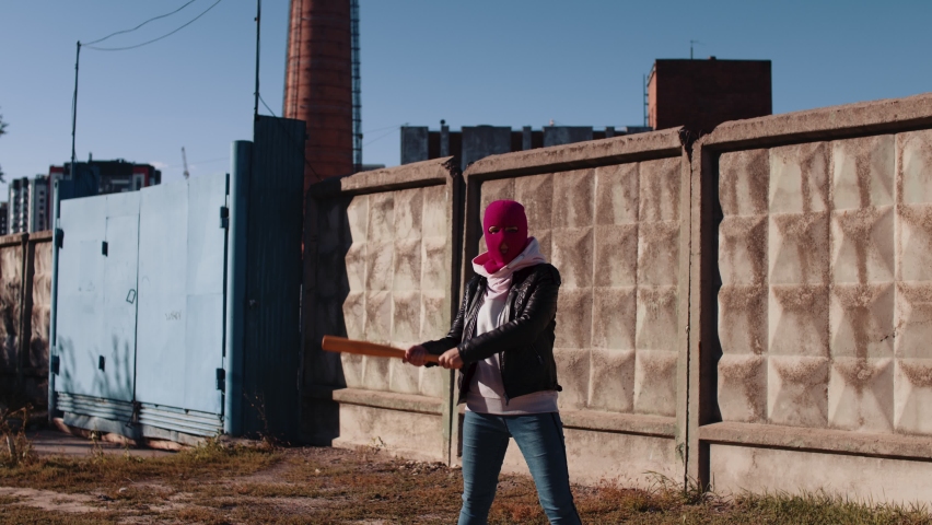 Young woman in pink balaclava with baseball bat in hands. Hooligan in mask beats with bat, looking at camera.
