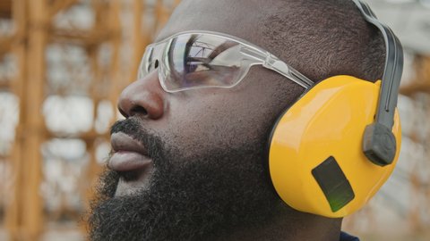 Extreme close up of black male worker in safety goggles putting on noise-canceling earmuffs at construction site