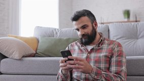 Handsome man using smartphone sitting at home. Happy smiling young man using mobile phone apps, texting message, browsing internet, watching video,tapping,looking at smartphone.