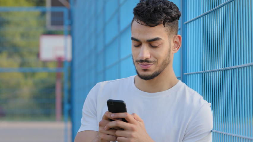 Young Middle eastern Arabic athlete in summer sportswear holding mobile phone, using virtual app, online service on smartphone, texting, chatting typing message, touching screen, standing at stadium | Shutterstock HD Video #1079961986