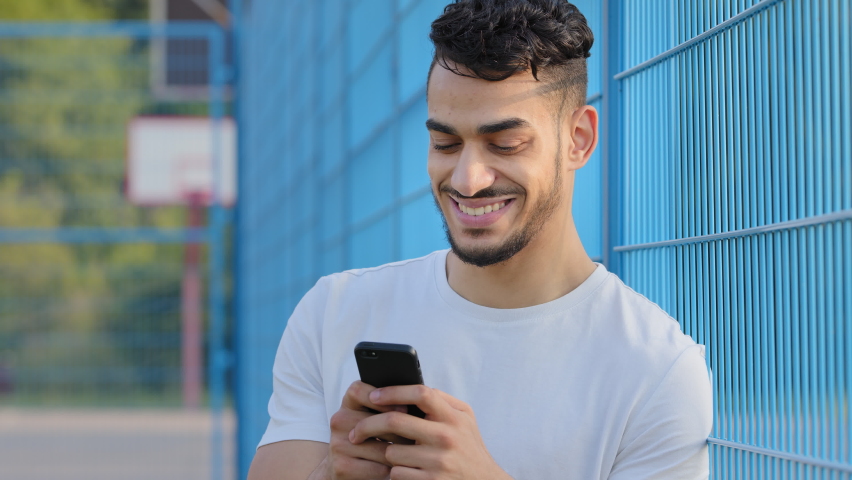 Young Middle eastern Arabic athlete in summer sportswear holding mobile phone, using virtual app, online service on smartphone, texting, chatting typing message, touching screen, standing at stadium | Shutterstock HD Video #1079961986