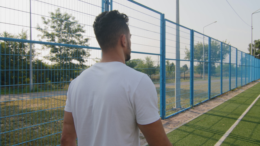 Back view Young Middle eastern Arabic athlete in summer sportswear white t-shirt walking moving along stadium fence returns after long workout, run or soccer. Indian runner finished training or game Royalty-Free Stock Footage #1079962085